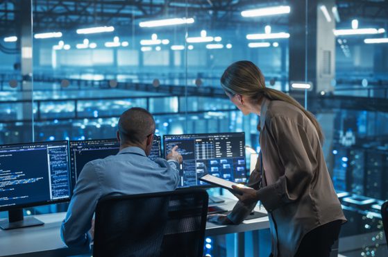 A woman discusses with a man in an office overlooking a data centre with multiple servers; the woman holds a tablet, and the man works on a computer with multiple screens.