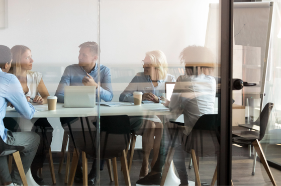 Several colleagues sit around a glass-walled meeting room table, with laptops and coffee cups, as a flipchart stands nearby.