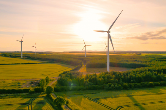 A wind farm with tall turbines in a green landscape during sunset, with golden sunlight filtering through the clouds and casting long shadows on the fields.