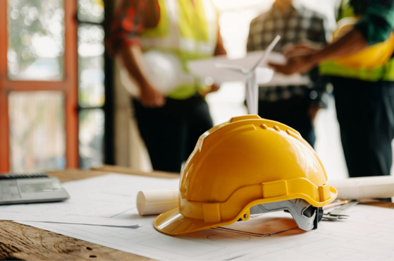 A yellow safety helmet placed on a wooden table with blueprints, with three people in high-visibility vests discussing plans in the background.