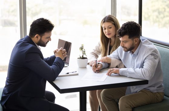 Three people sitting at a table in a bright office, engaged in a discussion, with papers, a pen, and a small potted plant on the table.