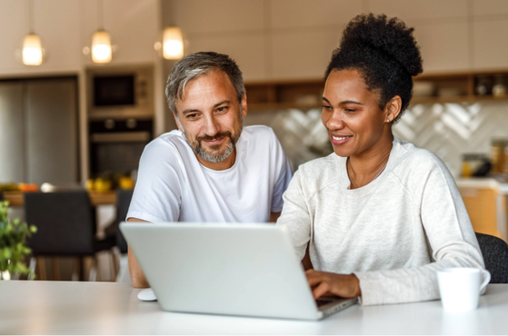 A man and woman sit at a table in a modern kitchen, looking at a laptop with smiles. The kitchen has warm lighting and wooden accents.