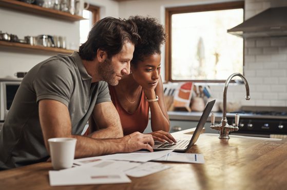 A man and woman sit at a kitchen counter, looking at a laptop with papers and a mug nearby, with a bright window in the background.