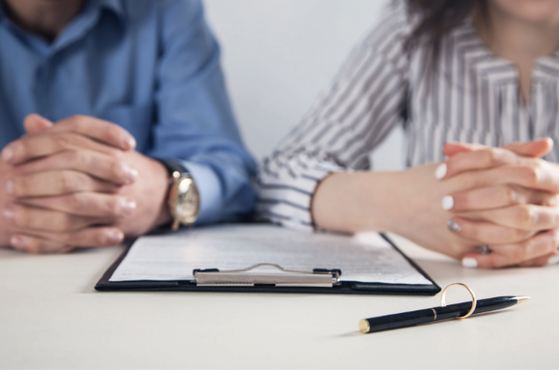 Two people sit at a white table with a clipboard and pen, hands folded in front, suggesting a formal meeting or interview in a bright, professional setting.