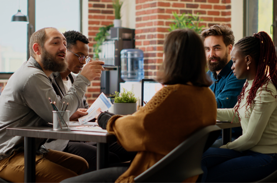 Five diverse colleagues sit around a table in a bright open-plan office, sharing documents and laptops while discussing; brick wall, plant and water cooler behind them.