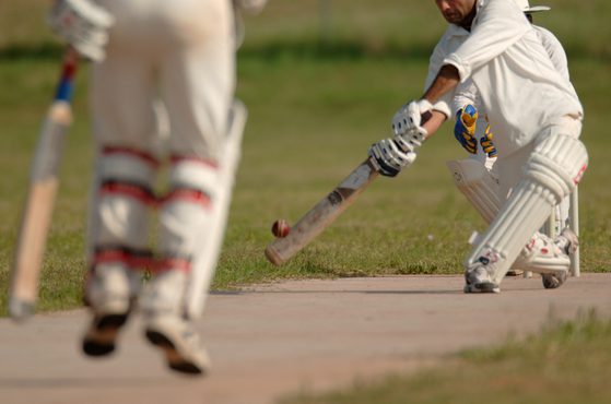 A cricket player in white pads and gloves attempts a shot with a bat, while another player, also in white, stands nearby on a grassy field.