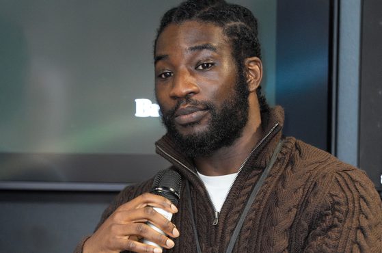 A man with dreadlocks and a beard sits on a chair, holding a microphone, wearing a brown cardigan and a smartwatch, with a dark background and a screen behind him.