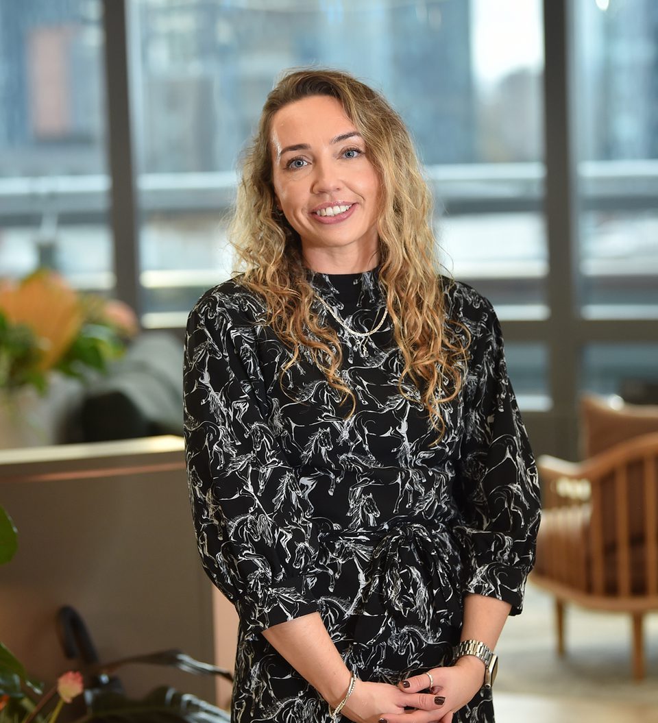 A woman with curly blonde hair smiling in a modern office with large windows, wearing a black dress with white abstract patterns, standing with hands clasped.