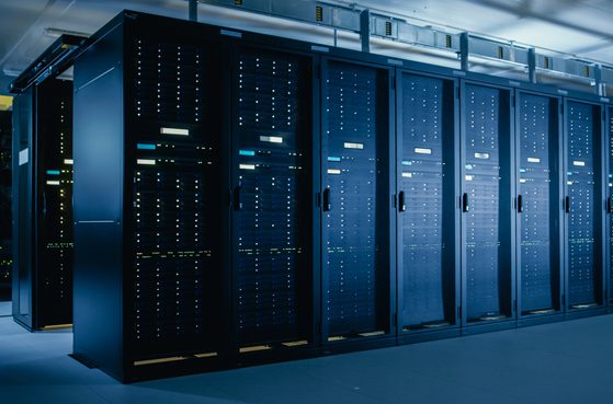 A row of black server racks with illuminated indicator lights in a data centre, with overhead cabling and cooling units visible.