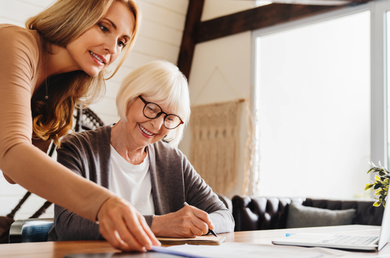 An elderly woman with white hair and glasses sits at a table, smiling and writing in a notebook while a younger woman with long red hair leans over, pointing at the notebook, in a bright room with large windows.