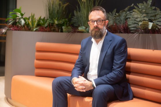Bearded person in a navy suit and white shirt sits on an orange cushioned bench indoors, hands clasped, with lush plants in the background.