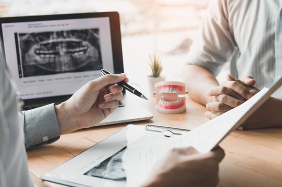 A dentist consultation with dental X-ray images on a computer screen, a dental model, and paperwork on a light wood table.