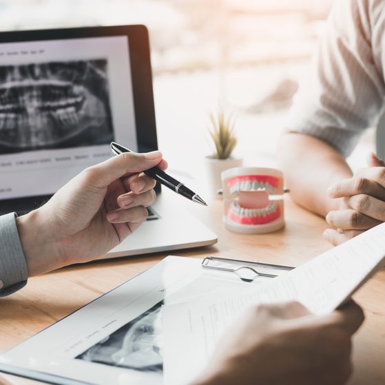 A dentist consultation with dental X-ray images on a computer screen, a dental model, and paperwork on a light wood table.