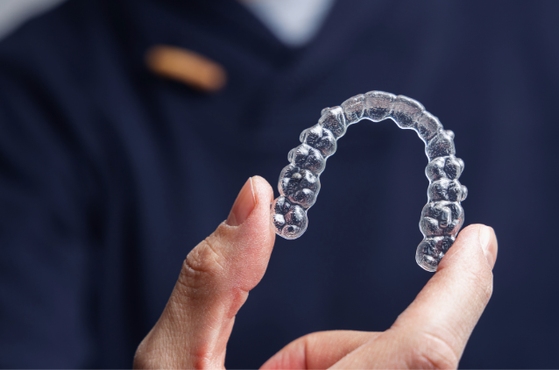 Clear dental aligner curved into an arch is held between fingertips against a dark blurred background.