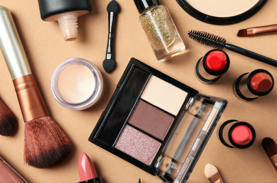 Assorted makeup items spread on a tan background, including a three-shade eyeshadow palette, brushes, nail polish, lipsticks, compact and mascara wand.