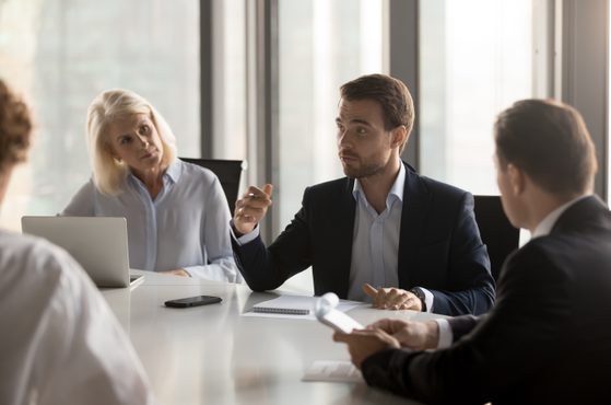 A group of four professionals in business attire engaged in a discussion in a modern office with large windows.