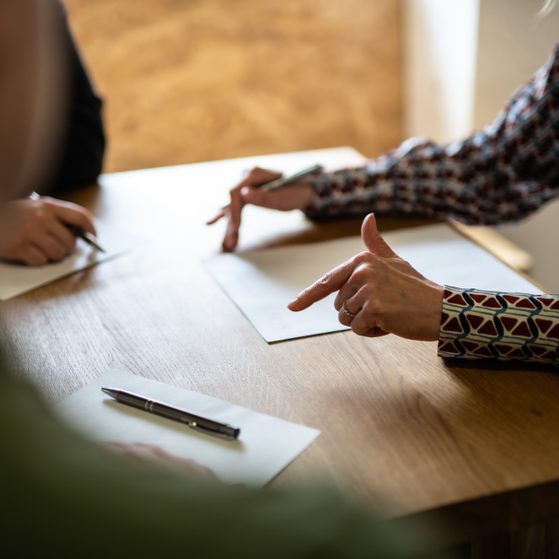 A person in a patterned long-sleeve shirt gesturing while sitting at a wooden table with papers and pens, engaged in a discussion with others.