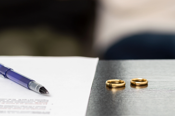Blue ballpoint pen on white paper sits beside two gold wedding rings on a dark wooden table, with a softly blurred background suggesting a contract or ceremony.