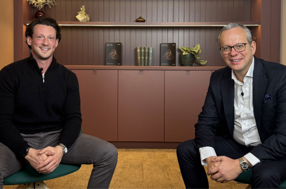Two men sit on chairs in a room with a wooden shelf holding books and decorations behind them. The man on the left wears a black sweater and grey pants. The man on the right wears a dark suit, white shirt, and glasses.