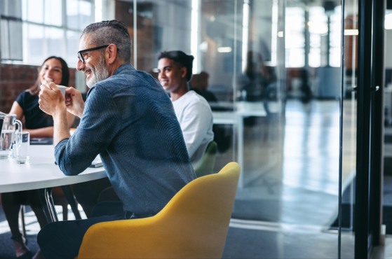 People in a modern office sit around a white table; a bearded person with glasses chats while others listen, with glass walls and a yellow chair in the foreground.