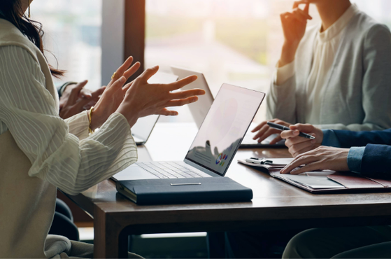 Three people sit around a table in a bright room, engaging in a discussion with laptops, papers, and pens, near a large window with natural light.