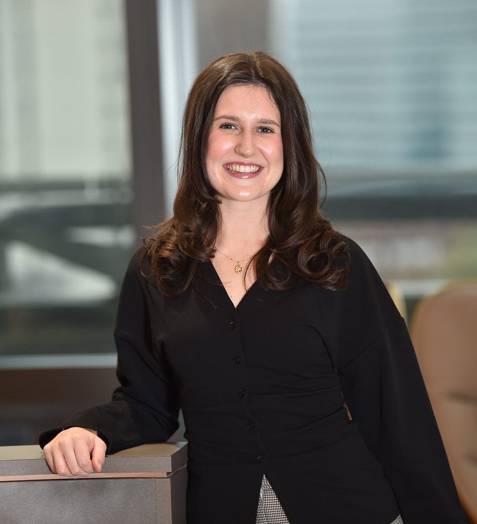 A woman with long, wavy brown hair wearing a black blouse, smiling, standing indoors near a desk or counter, with a modern office background.