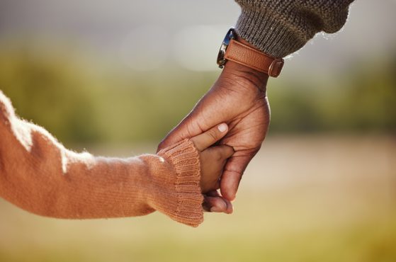 Close-up of an adult and child holding hands outdoors, with the adult wearing a brown leather watch strap and the child in an orange sweater, blurred background.