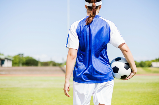 Back view of a player on a sunny football pitch, wearing a blue and white jersey with white shorts, holding a black-and-white football at their side.