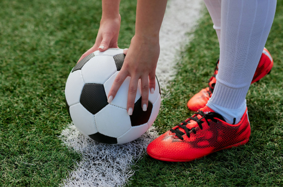 Person in football gear prepares to kick a black and white soccer ball on green grass, wearing white socks and bright red football boots.