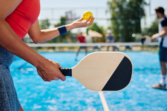 A person holds a padel racket near a court with a blue surface, with a yellow ball in one hand, while others are in the background.
