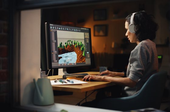 A woman with curly dark hair wearing headphones works on a computer with a 3D landscape design software, featuring trees, a river, and textured terrain.