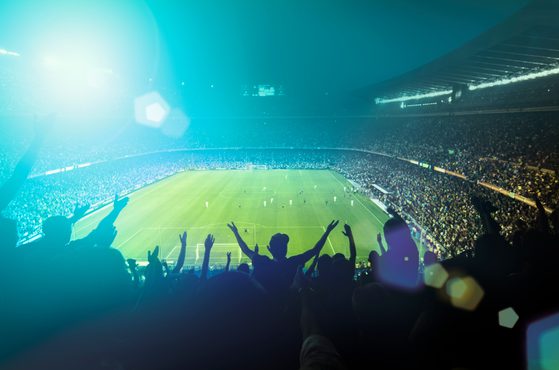 A packed football stadium with fans celebrating and raising their hands, illuminated by bright stadium lights, during a match under a dark sky.
