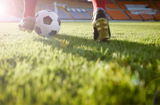 Soccer ball on a sunlit grass field with players' feet nearby, lens flare and an empty stadium in the background.
