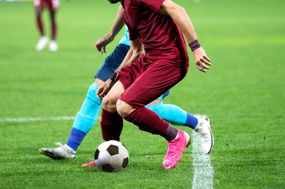 Players in soccer jerseys compete for the ball on a green field during a match, with some players in light blue and one in maroon, wearing pink boots.
