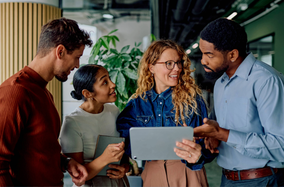Four diverse professionals are engaged in a discussion around a tablet in a modern office setting, with a large plant and green accent wall in the background.