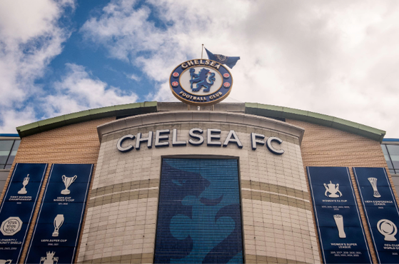 Chelsea FC stadium facade with a large CHELSEA FC sign, circular club crest atop, and trophy banners on either side against a blue sky.