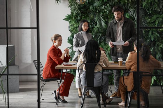 Five people are gathered around a table in a meeting room with a green leafy wall in the background. Four women are seated, and one man is standing, holding papers.