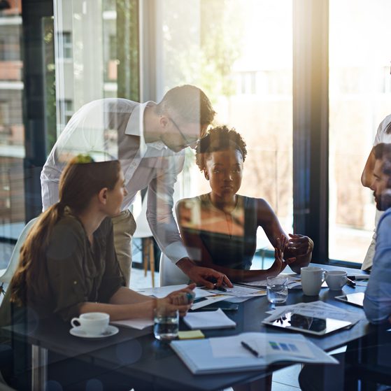 Six people in a business meeting around a table in a modern office with large windows and natural light.
