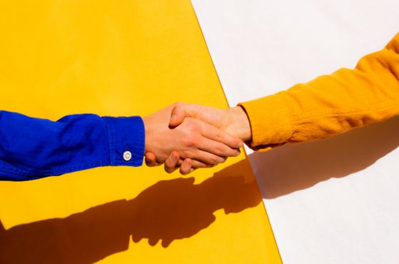 Two people shake hands, one in a blue sleeve and the other in a yellow sleeve, against a bold yellow and white split background with strong shadows.