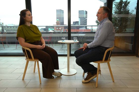 Two people are seated across from each other at a small round table, having a conversation in a modern indoor space with large windows and a cityscape view.