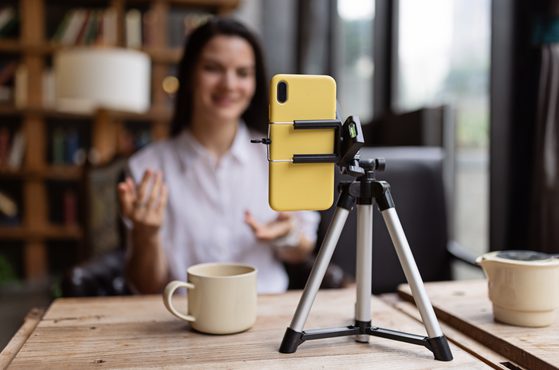 A smiling woman in a white shirt sits at a wooden table, with a yellow smartphone mounted on a tripod in front of her, camera aimed at her, in a cozy room with bookshelves.