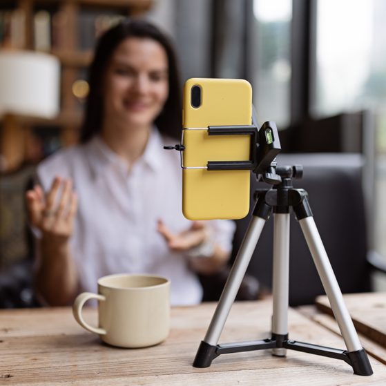 A smiling woman in a white shirt sits at a wooden table, with a yellow smartphone mounted on a tripod in front of her, camera aimed at her, in a cozy room with bookshelves.
