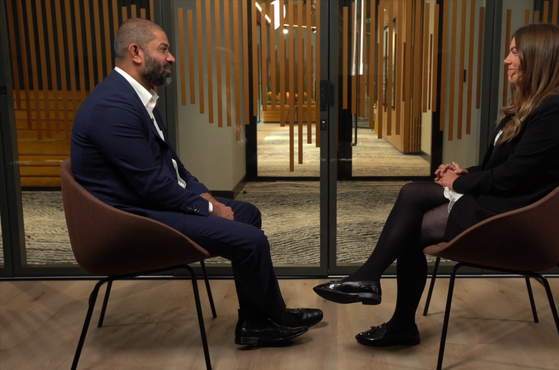 Two people sit facing each other in a modern office, separated by a glass door with wood slats in the background. The man on the left wears a navy suit, and the woman on the right wears a black blazer.