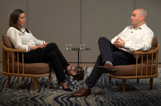 A woman and a man sit facing each other on separate armchairs, with a small table holding two glasses between them in a neutral-toned room.