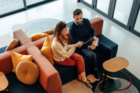 A man and woman sit on a Couch with colourful cushions and look at a tablet together in a modern, glass-walled lounge area with a grey circular rug.