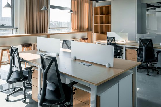 An empty modern office with white desks separated by small partition screens, black ergonomic chairs, large windows with beige curtains, and wooden shelves.