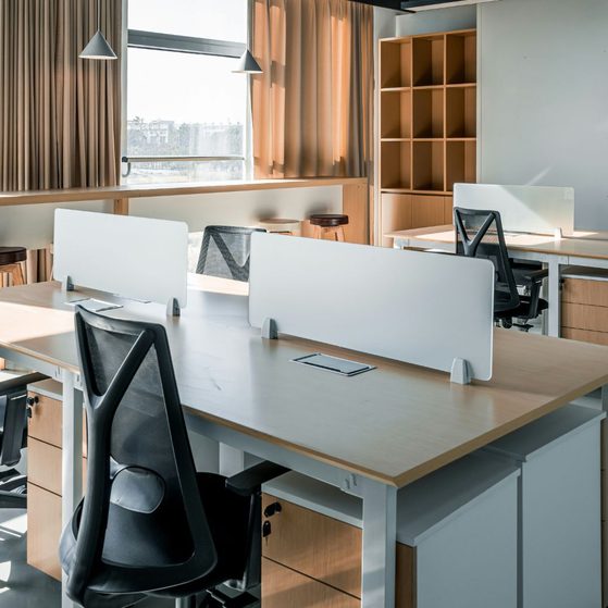An empty modern office with white desks separated by small partition screens, black ergonomic chairs, large windows with beige curtains, and wooden shelves.