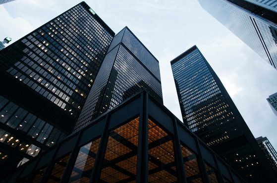Several tall modern glass skyscrapers viewed from below, with reflections on their surfaces and a partly cloudy sky in the background.
