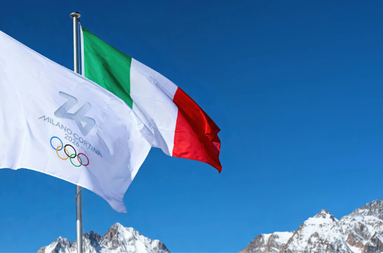 Three flags flying against a backdrop of snow-capped mountains and a clear blue sky: an Italian flag, a white flag with Milano Cortina 2026 Olympic logo, and an Olympic flag.
