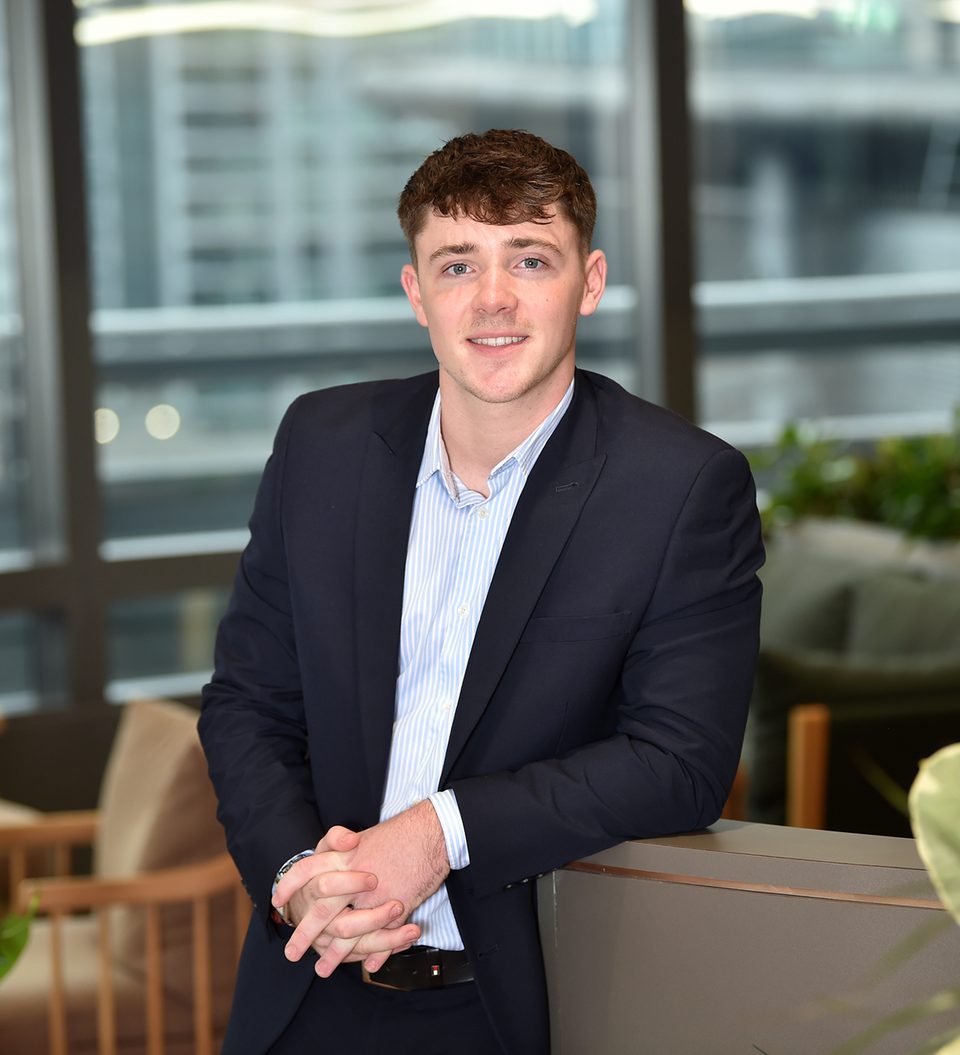 A young man in a dark suit and light blue shirt, smiling, leaning on a grey partition in a modern office space with large windows and blurred cityscape background.